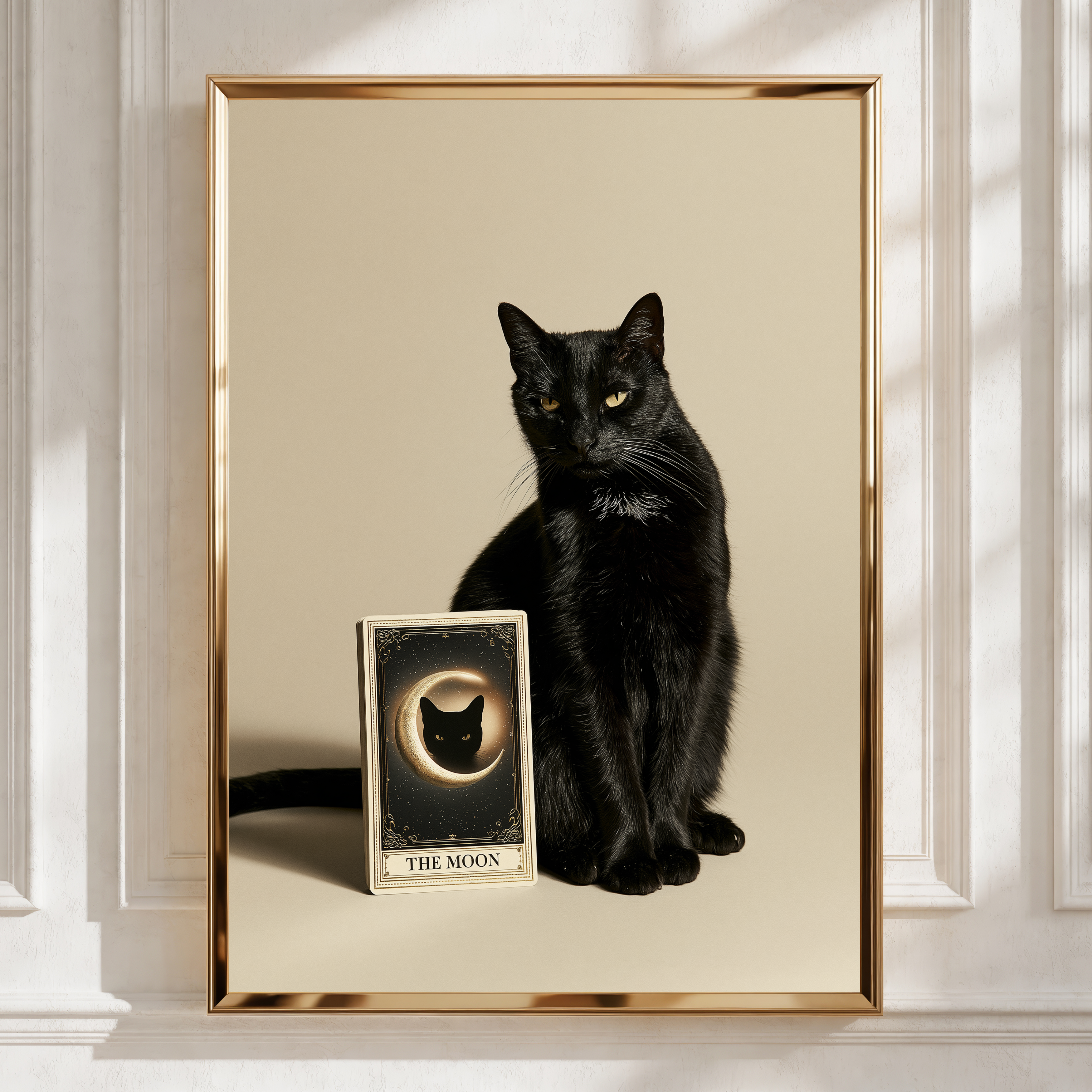 A framed art print featuring a black cat sitting beside a tarot card labeled 'THE MOON' on a warm cream background, enclosed in a thin gold frame and displayed against a white paneled wall.