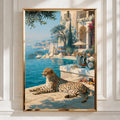 A framed vertical art print depicting a cheetah resting by a blue sea beside a luxurious coastal villa with a pool, displayed on a white paneled wall.