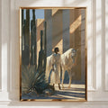 A framed art print featuring a desert scene with a white horse, a person in a long white robe, and tall cacti, displayed on a white paneled wall with soft window shadows.