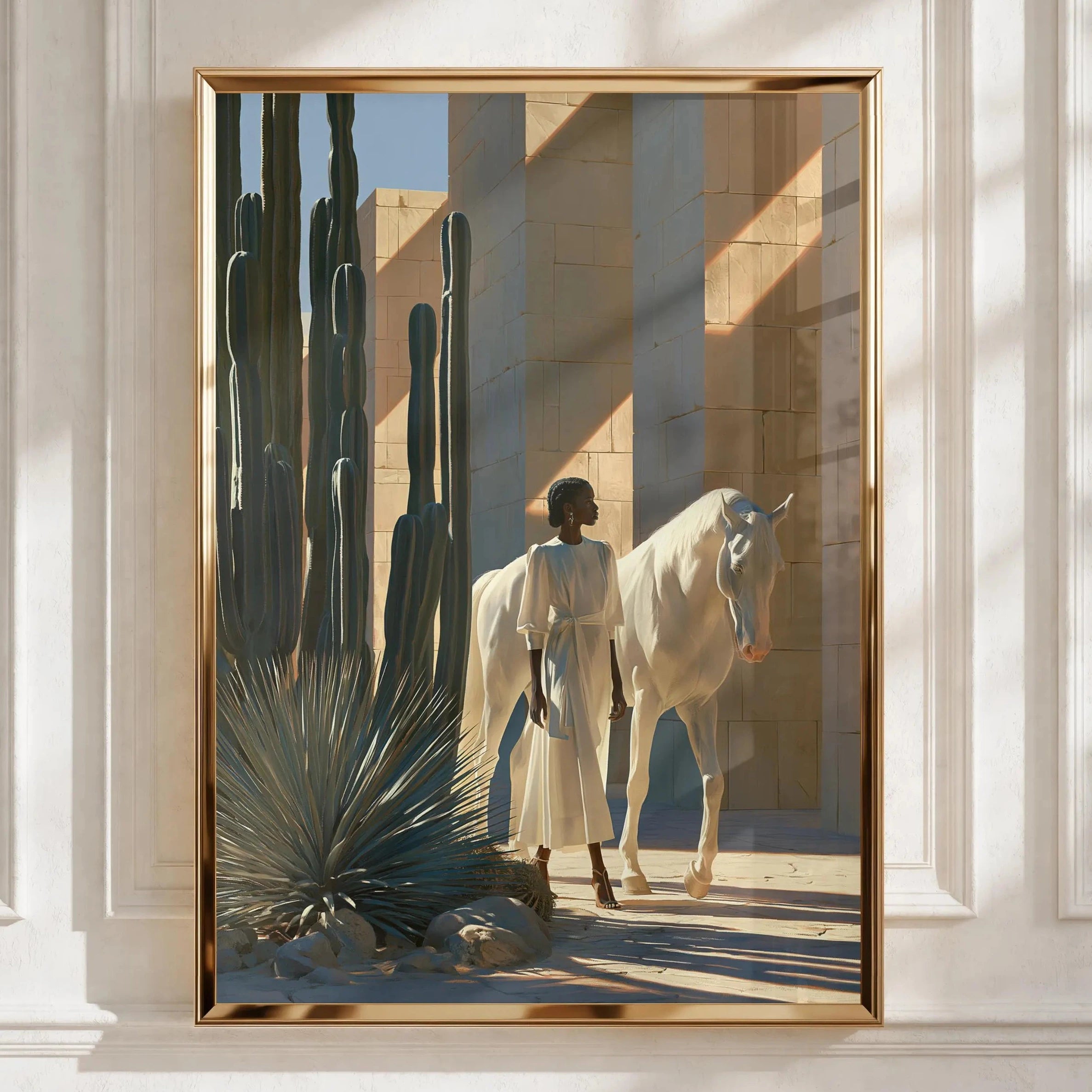 A framed art print featuring a desert scene with a white horse, a person in a long white robe, and tall cacti, displayed on a white paneled wall with soft window shadows.